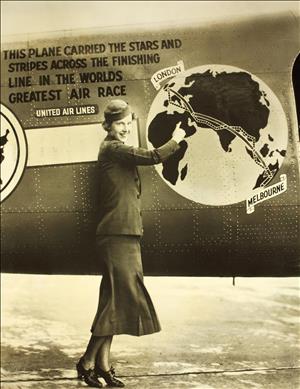 A smiling woman in a hat points to a map painted on the side of an airplane